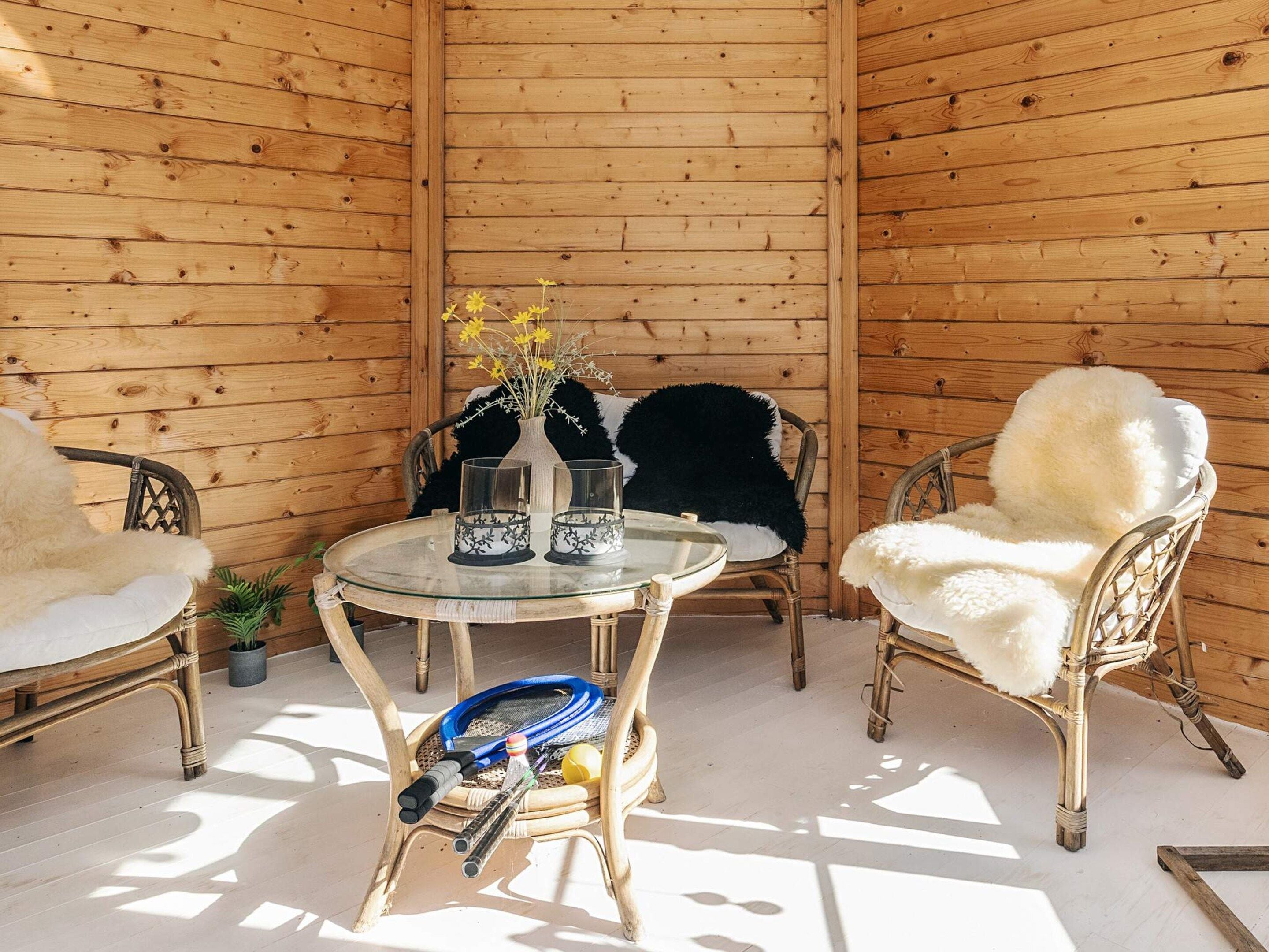 A wooden-paneled room with a round glass table, two wicker chairs with white fur throws, and a vase with yellow flowers.