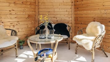 A wooden-paneled room with a round glass table, two wicker chairs with white fur throws, and a vase with yellow flowers.