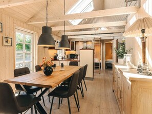 A spacious kitchen with wooden cabinets, a central island, pendant lighting, and a view through an open door to a bedroom. - 4 Star Holiday Home in Vaeggerlose (Vaeggerlose)