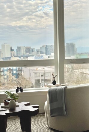 Ein Zimmer mit einem großen Fenster, einem Sessel, einem kleinen Tisch mit einer Pflanze und einer Vase.