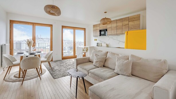 Ett modernt vardagsrum med en soffa, ett matbord och stolar, ett kök med träskåp och stora fönster med utsikt över staden.
