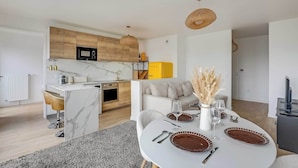 Ett modernt kök med träskåp, en mikrovågsugn och en gul köksapparat. Det finns en matplats med ett vitt bord dukat för två personer, en vas med torkade växter och en soffa i bakgrunden.