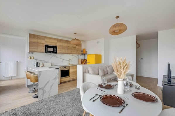 A modern kitchen with wooden cabinets, a microwave, and a yellow appliance. There is a dining area with a white table set for two, a vase with dried plants, and a sofa in the background.