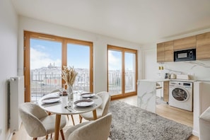 A modern kitchen with a built-in microwave and washing machine, large windows with a city view, and a dining area with a table set for two.