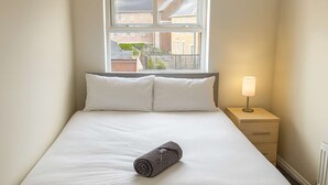 Ein ordentlich gemachtes Bett mit weißer Bettwäsche und einem zusammengerollten Handtuch, ein Nachttisch mit Lampe und ein Fenster mit Blick auf Gebäude.