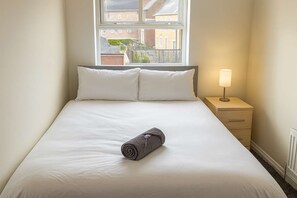 Ein ordentlich gemachtes Bett mit weißer Bettwäsche und einem zusammengerollten Handtuch, ein Nachttisch mit Lampe und ein Fenster mit Blick auf Gebäude.