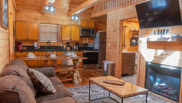 A cozy cabin interior with a living room, wooden dining table, kitchen area, and a fireplace. - Lazy Bear Den | Cozy w/ Hot Tub (Sevier County)