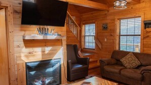 A cozy log cabin interior with a fireplace, a television mounted on the wall, a comfortable sofa, and a wooden coffee table. - Lazy Bear Den | Cozy w/ Hot Tub (Sevier County)