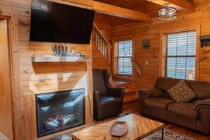 Ein gemütliches Blockhaus-Interieur mit Kamin, einem an der Wand montierten Fernseher, einer bequemen Couch und einem Holztisch.