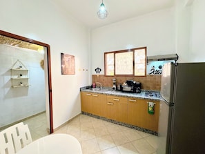 A compact kitchen with a refrigerator, stove, and sink. There is a window with decorative glass, a small shelf with items, and a door leading to another room. - Jnane awrad house e relaxation and authenticity (Marrakech)