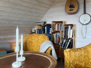 Een gezellige kamer met een houten plafond, een ronde houten tafel, twee stoelen met gestreept zitoppervlak, een boekenplank vol boeken en een banjo aan de muur.