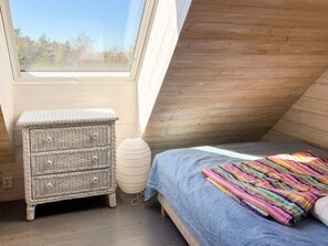 Een kamer met een schuine kap, een bed met een blauwe deken, een rieten nachtkastje en een witte lamp.