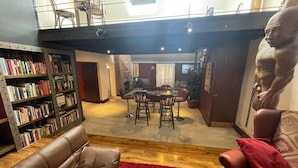 Una sala de estar con un sofá de cuero, un tapete rojo, una zona de comedor con mesa y sillas, y una estantería llena de libros.