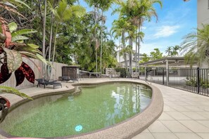 Ein Pool mit geschwungener Kante, umgeben von einem Zaun, neben einem Gebäude mit einer überdachten Terrasse.