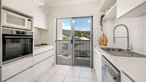 A modern kitchen with a built-in oven, microwave, and dishwasher, a sliding glass door to a balcony, and a stainless steel sink. - Trinity Beach Bliss: Experience Tropical Living (Trinity Beach)