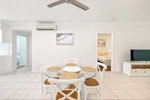 Un salon moderne avec une table ronde en verre, des chaises blanches, un ventilateur de plafond et un téléviseur posé sur un meuble blanc.