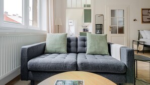Ein graues Sofa mit zwei gemusterten Kissen, einem Holztisch und im Hintergrund Blick auf eine Küche und ein Bett.