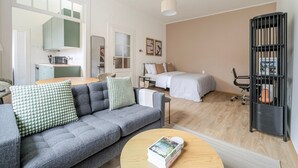 Ein modernes Wohnzimmer mit einem grauen Sofa, einem runden Couchtisch, einem Bett mit weißer Bettdecke, einer schwarzen Bücherwand und einer Küchenzeile inklusive Mikrowelle.