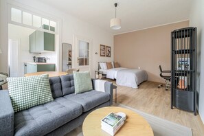 Ein modernes Wohnzimmer mit einem grauen Sofa, einem runden Couchtisch, einem Bett mit weißer Bettdecke, einer schwarzen Bücherwand und einer Küchenzeile inklusive Mikrowelle.