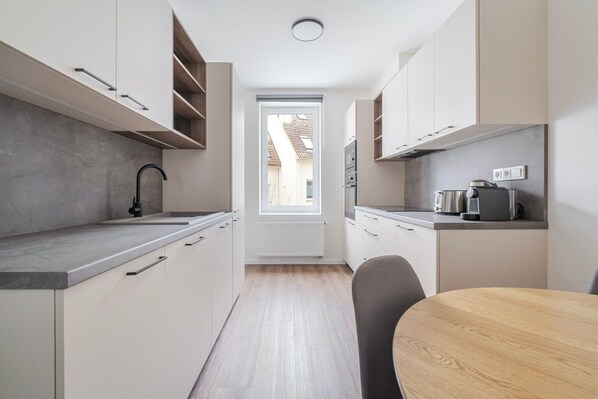 A narrow kitchen with white cabinets, a grey countertop, a black faucet, and a window with a view of a building outside. - Blueground | Žižkov, view, nr Vitkov Hill (PRG-96) (Prague)