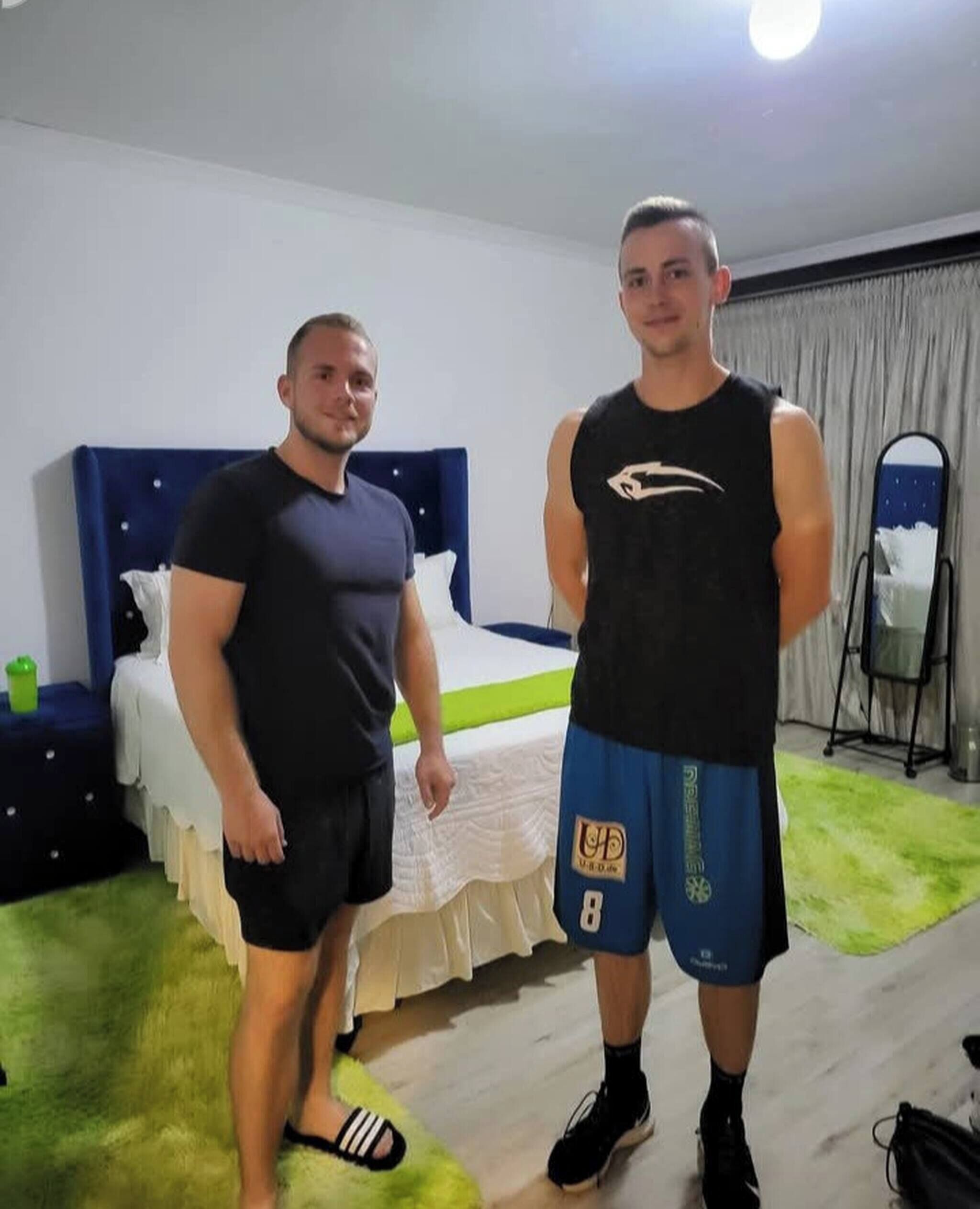Two men in a room with a bed, a nightstand, and a mirror.