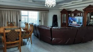 A living room with a brown leather sofa, a wooden dining table with chairs, a television, and a chandelier.