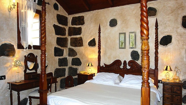 A four-poster bed with a stone wall backdrop, a wooden nightstand with a lamp, and a mirror on the left side. - Holiday Home in Vera de Erques (Guia de Isora)