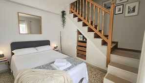 A bedroom with a bed, a nightstand, a mirror, a staircase, and framed pictures on the wall. - Maisonette With Beautiful Terrace (Mykonos)