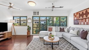 A modern living room with a sectional sofa, a flat-screen TV, a ceiling fan, and large windows with blinds.