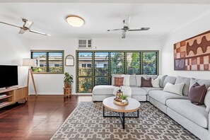 A modern living room with a sectional sofa, a flat-screen TV, a ceiling fan, and large windows with blinds.