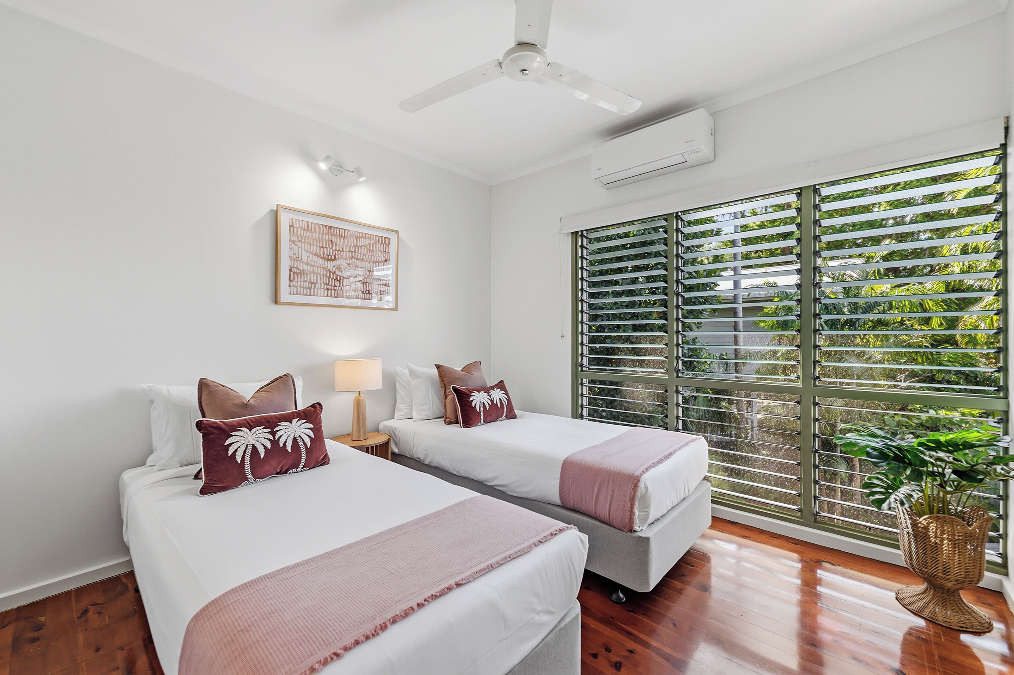 Une chambre d’hôtel avec deux lits, un ventilateur de plafond, un appareil de climatisation, une fenêtre avec des stores et un tableau encadré au mur.