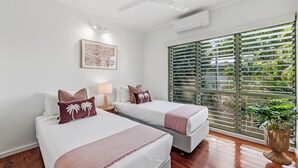 A hotel room with two beds, a ceiling fan, an air conditioning unit, a window with blinds, and a framed picture on the wall.