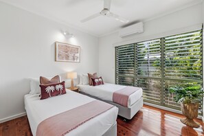 A hotel room with two beds, a ceiling fan, an air conditioning unit, a window with blinds, and a framed picture on the wall.