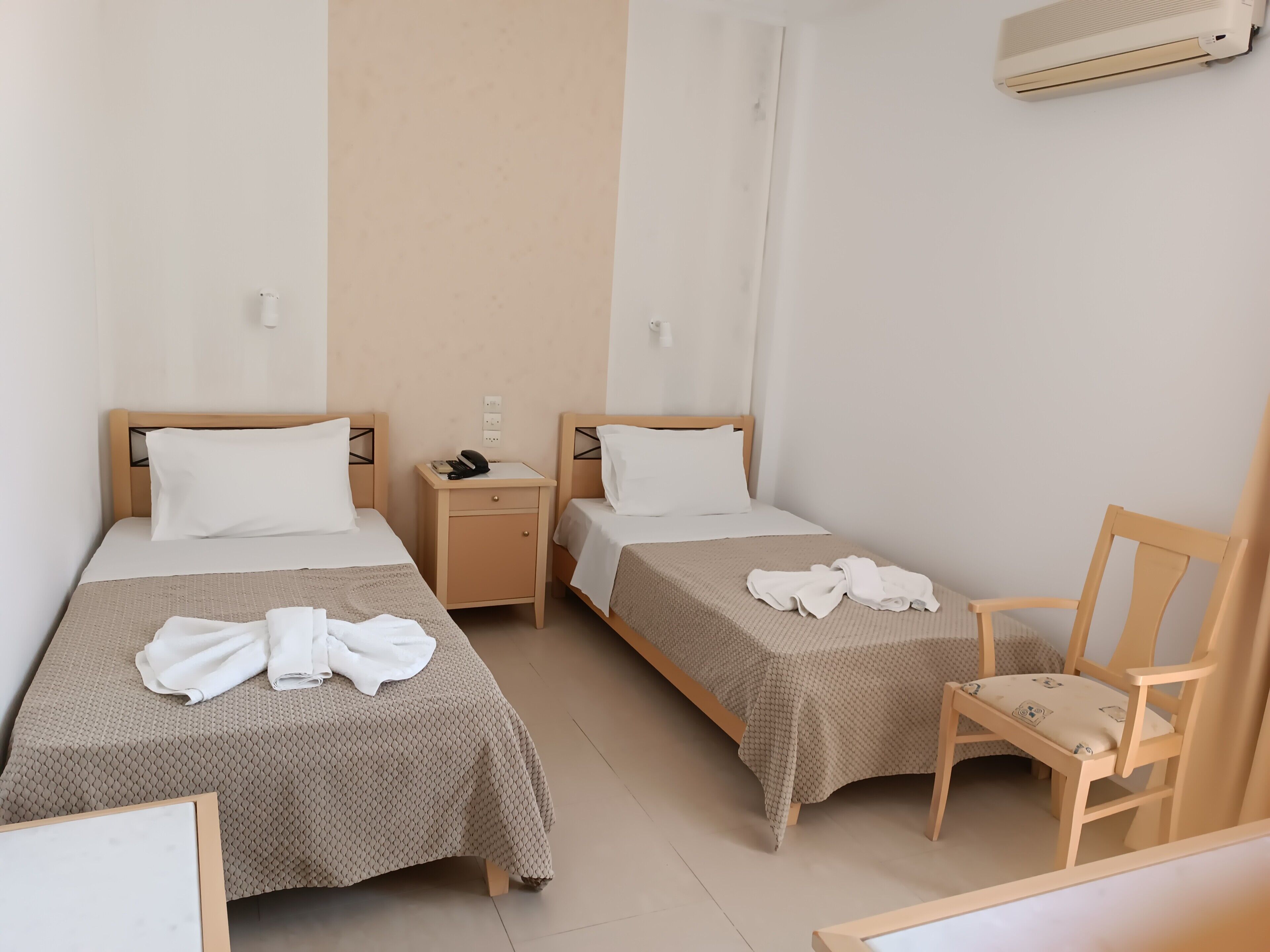 A hotel room with two single beds, a wooden nightstand, a chair, and an air conditioning unit.