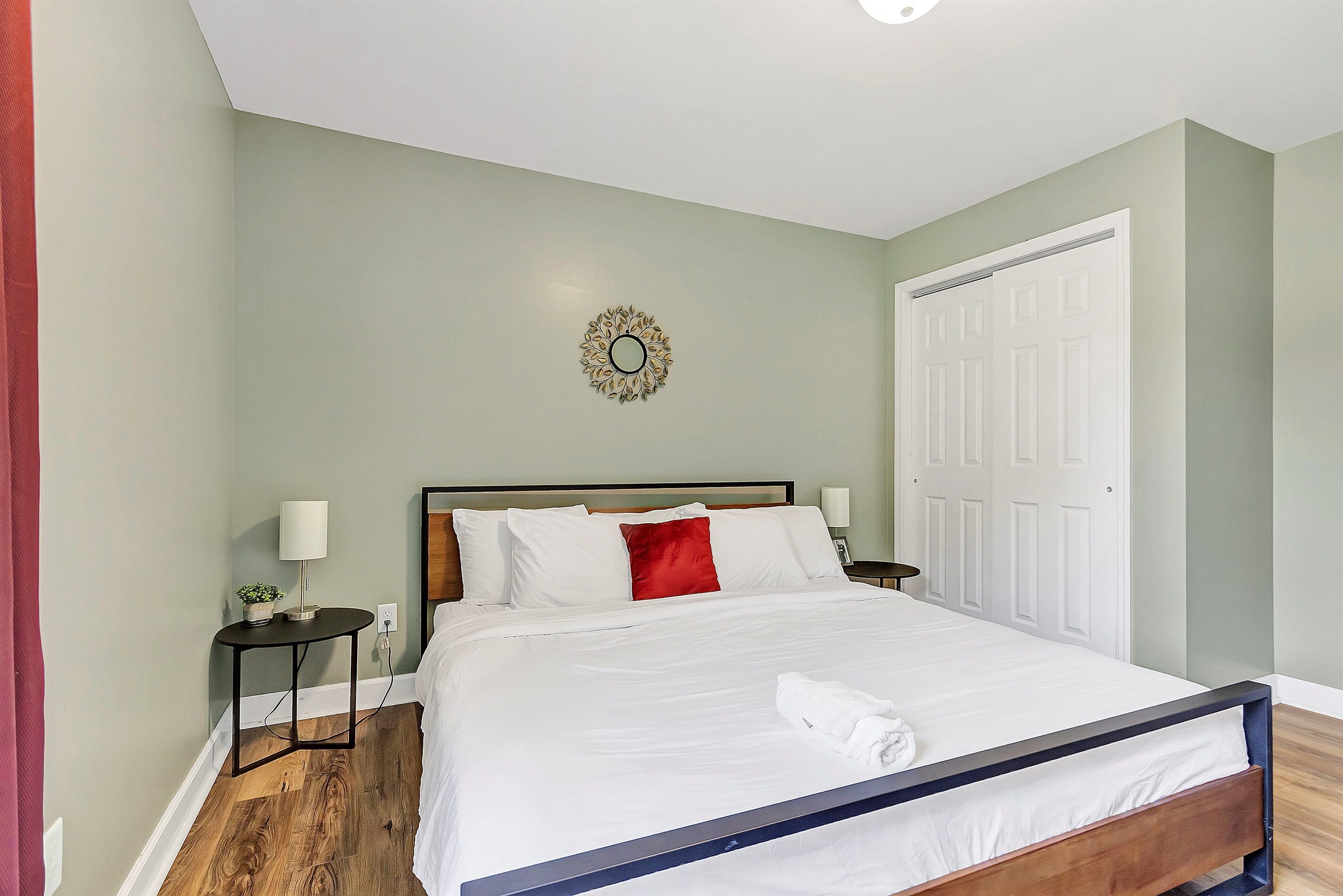 Un lit bien fait, avec du linge de lit blanc et un coussin rouge, une petite table de chevet avec une lampe, et un objet décoratif fixé au mur.