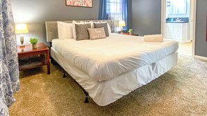 Un dormitorio con una cama grande, dos mesitas de noche, una mesita auxiliar, una ventana con cortinas y una puerta que da a un baño.