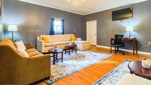 Una sala de estar con sofá, sillón, mesa de centro, alfombra, candelabro y televisor de pantalla plana montado en la pared.