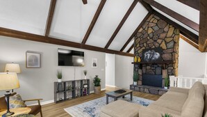 A living room with a stone fireplace, wooden beams, a sofa, a coffee table, and a TV mounted on the wall. - Lake View Landing | Pool/Groups/Nature/Games (Birmingham)