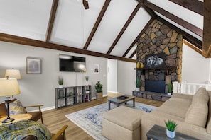 A living room with a stone fireplace, wooden beams, a sofa, a coffee table, and a TV mounted on the wall. - Lake View Landing | Pool/Groups/Nature/Games (Birmingham)