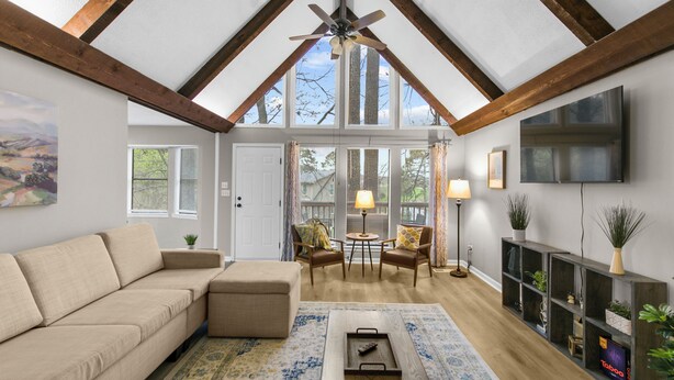 A spacious living room with a high ceiling, wooden beams, a large window, a sofa, chairs, a coffee table, and a TV. - Lake View Landing | Pool/Groups/Nature/Games (Birmingham)