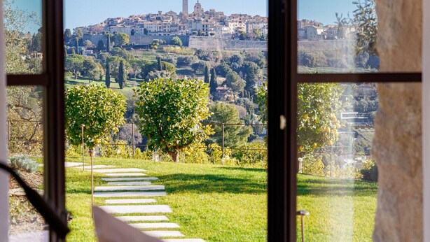 Blick durch ein Fenster auf einen gepflasterten Weg, der zu einer Grünfläche führt, ein Gebäude in der Ferne und ein weißes Kissen im Vordergrund.