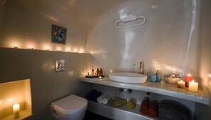 A bathroom with a white sink, toilet, and shelves with towels and toiletries. - Fira Home With Rooftop Hot Tub (Santorini)