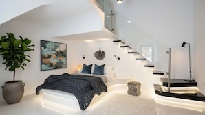 A modern bedroom with a bed, bedside table, lamp, and a large potted plant. - Fira Home With Rooftop Hot Tub (Santorini)