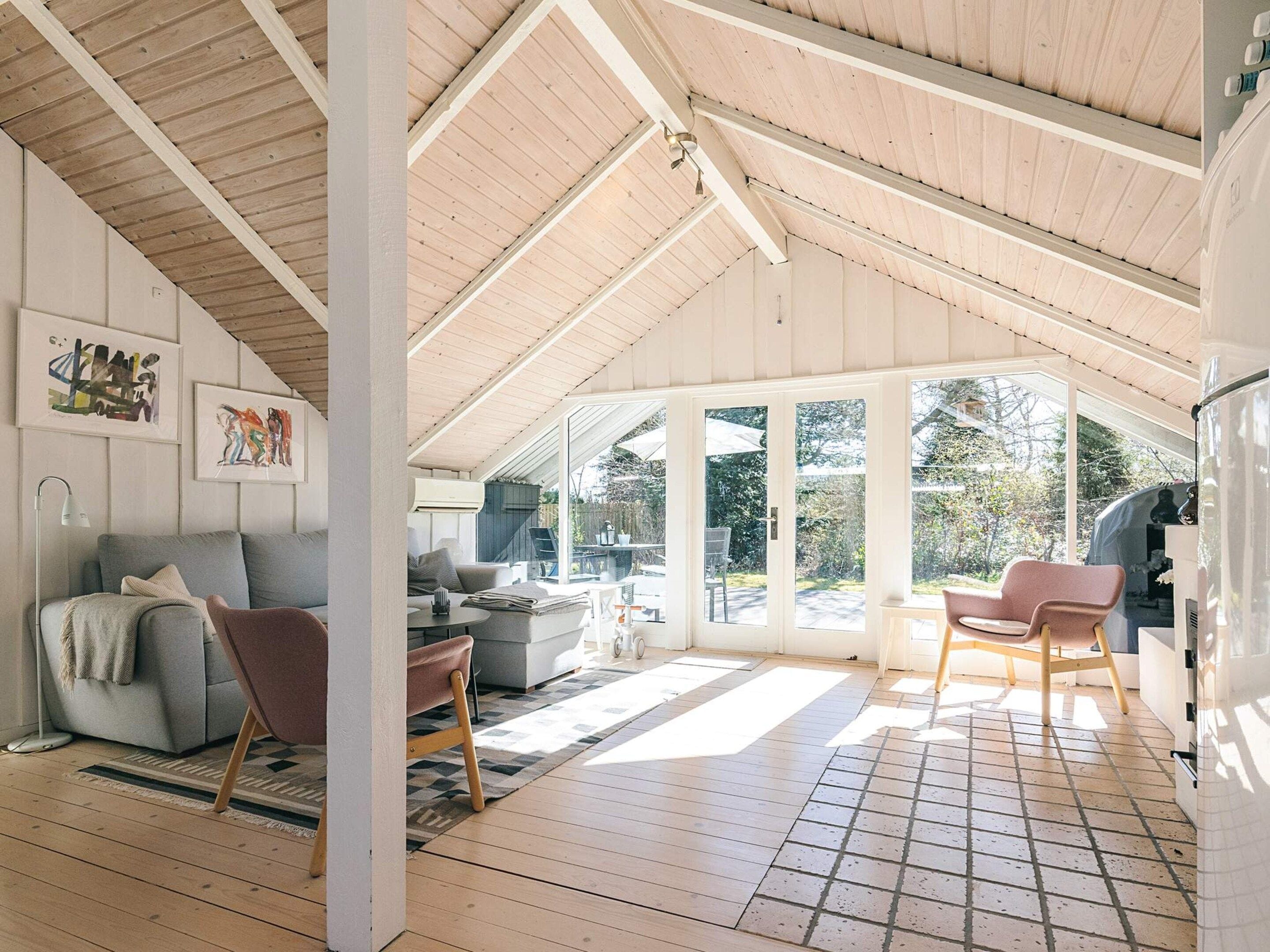 A spacious room with a high ceiling, wooden beams, a large sliding glass door leading outside, a grey sofa, a pink chair, and a white refrigerator.