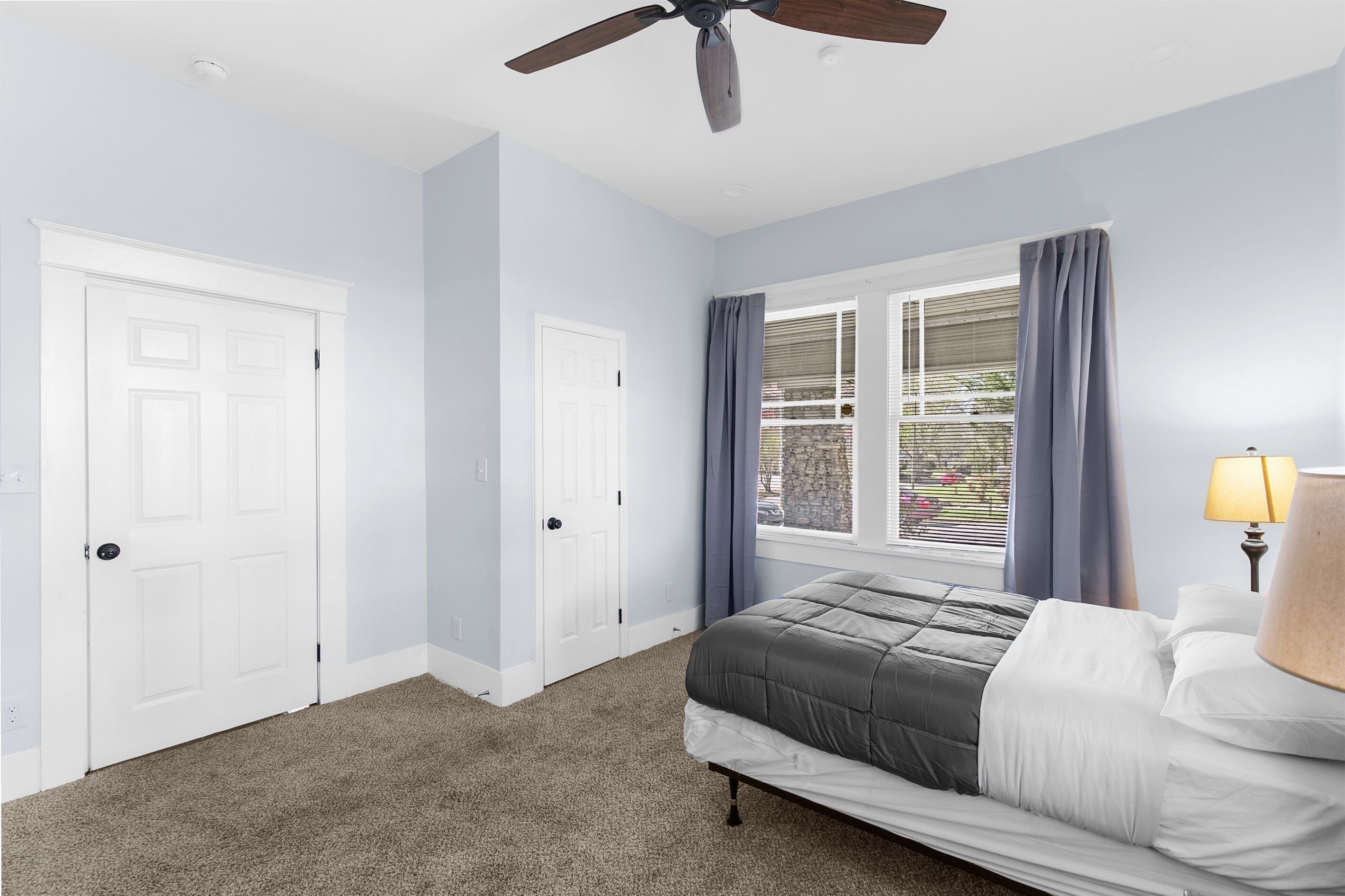 Un dormitorio con cama, ventilador de techo, ventana con cortinas y una puerta.