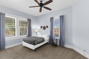 A bedroom with a bed, a ceiling fan, two windows with curtains, a nightstand with a lamp, and a wall-mounted clock.