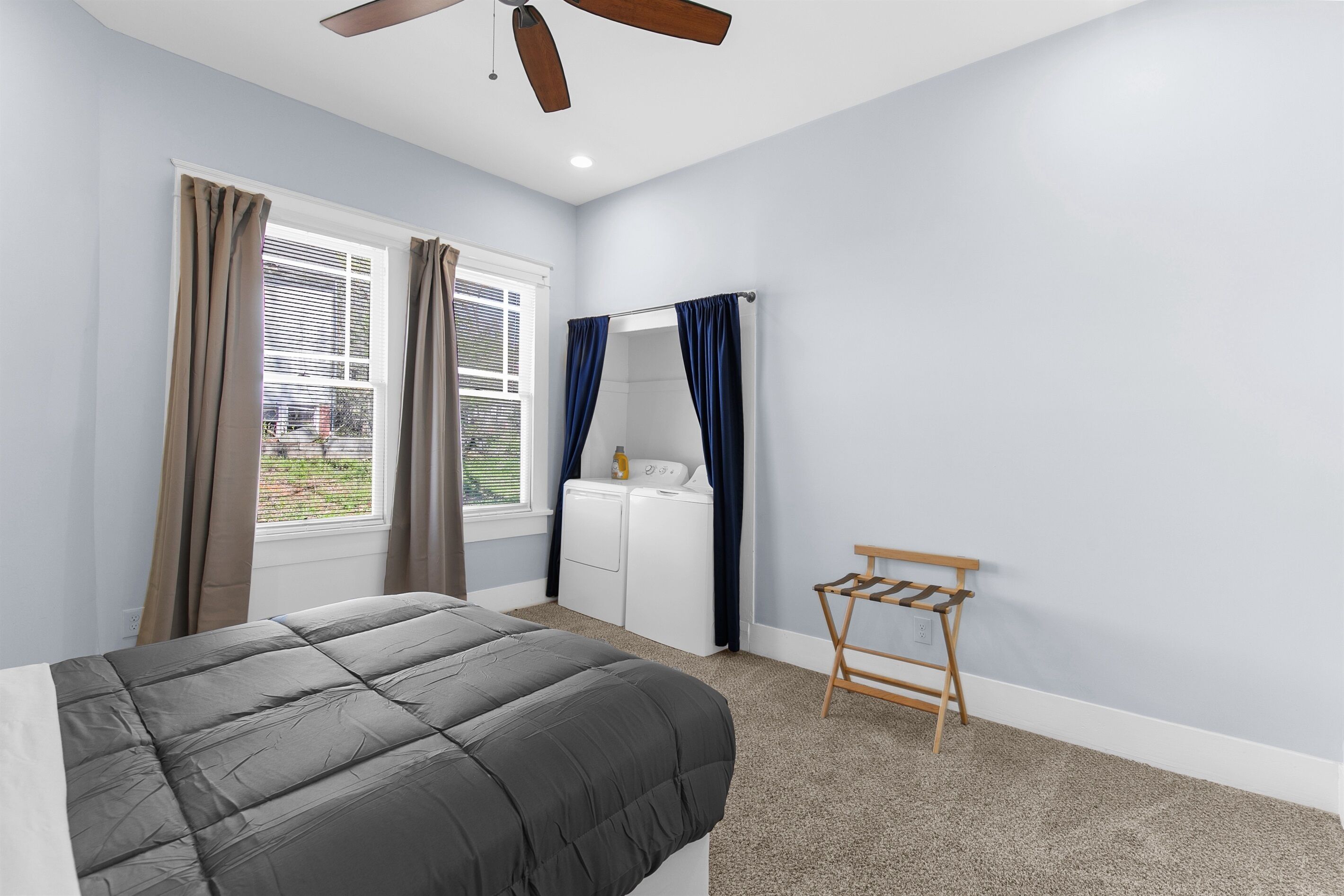 Un dormitorio con cama, ventilador de techo, ventana con cortinas, lavadora y un tendedero de madera.