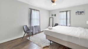 Un dormitorio con cama, una silla, ventilador de techo y dos ventanas con cortinas.