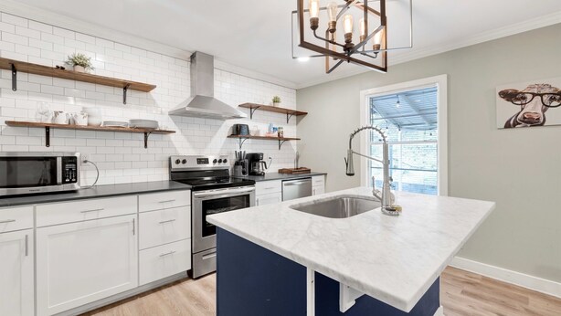 Una cocina moderna con armarios blancos, una isla central con pileta, estufa y microondas. Hay estantes de madera sobre la encimera y una lámpara colgante.