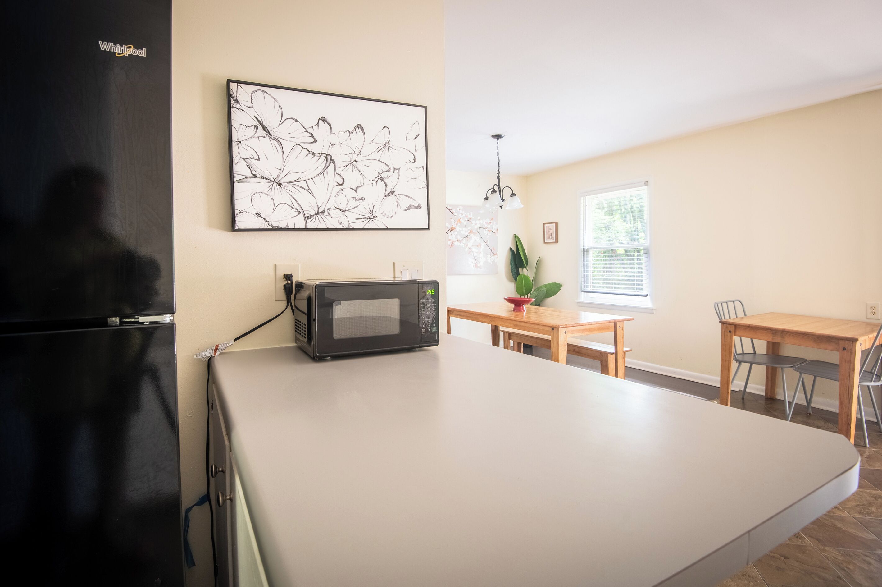 Una cocina con un refrigerador negro, un microondas, una mesa de comedor con sillas y una ventana con persianas.